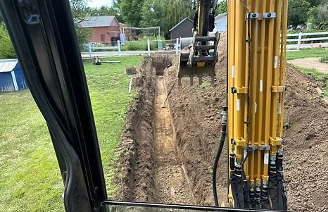 A yellow excavator digging a long trench in a grassy yard.