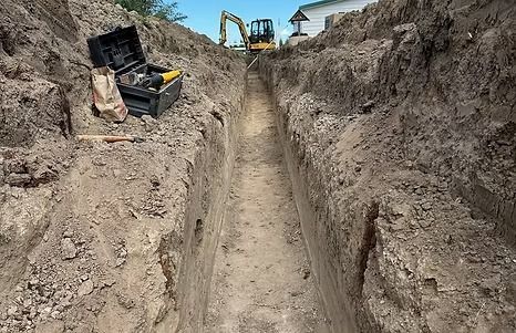 Long, narrow trench dug in dirt, construction site with excavator in background. Tools sit on the edge.