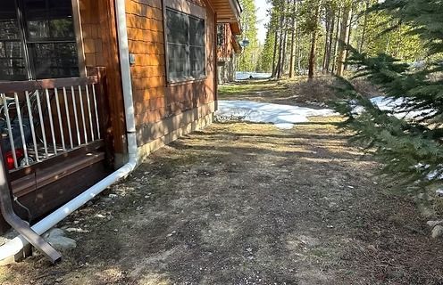 Side of a wood cabin with a brown facade, a pathway covered in dirt and patches of snow, and a forest.