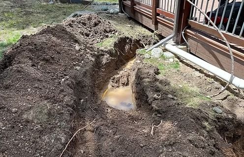 A trench dug in dirt near a wooden structure is filled with muddy water.