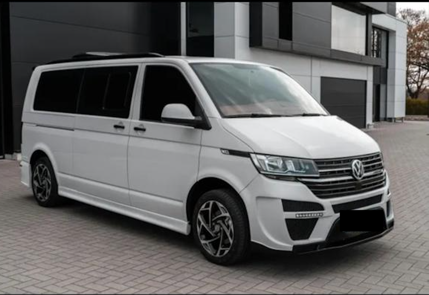 Biały van Volkswagen z czarnymi akcentami, zaparkowany przed nowoczesnym budynkiem.