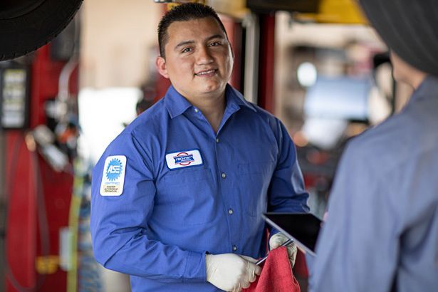 Mechanic in blue uniform smiles at a customer in a car repair shop, holding a rag and tablet.