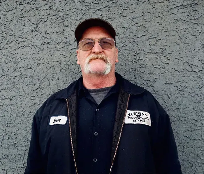 Man with mustache wearing a cap and work jacket, standing in front of a gray wall.