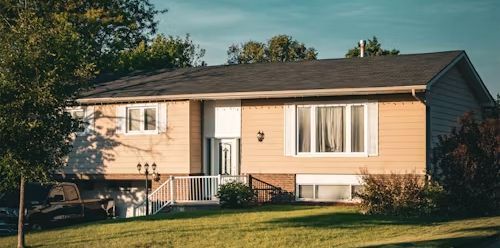 A two-story tan house with a black roof, green lawn, and trees.