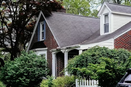 Two-story house with brick and white siding, dark gray roof, and white picket fence in front of greenery.