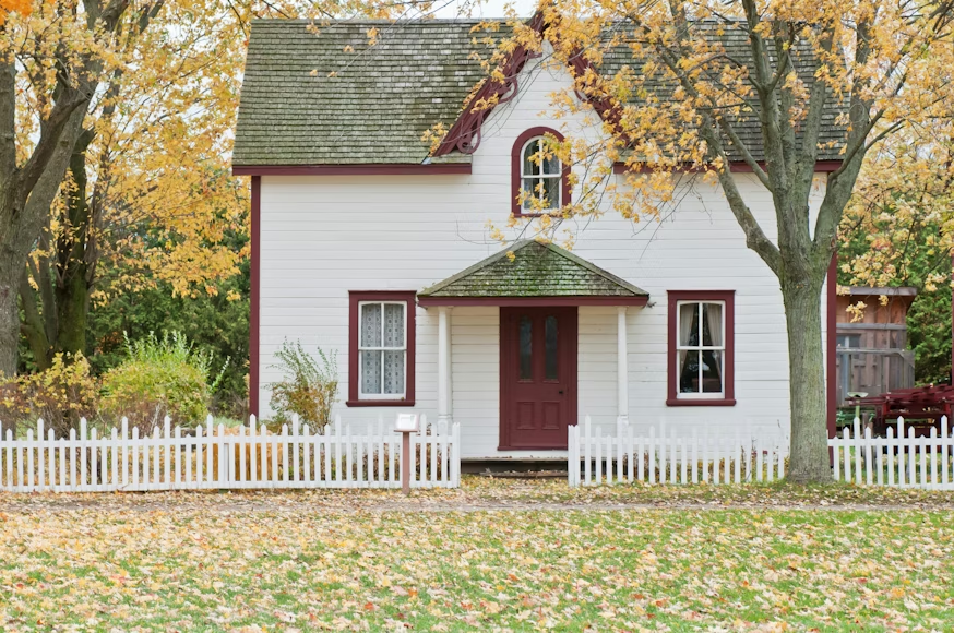 A white house with a maroon door and trim, surrounded by yellow autumn trees and a white picket fence.