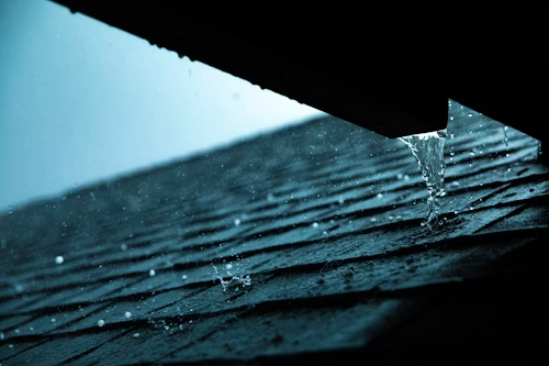 Raindrops falling from a roof edge onto dark shingles in blue light