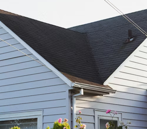 Gray house roof with black shingles, white siding, and pink flowers in the foreground