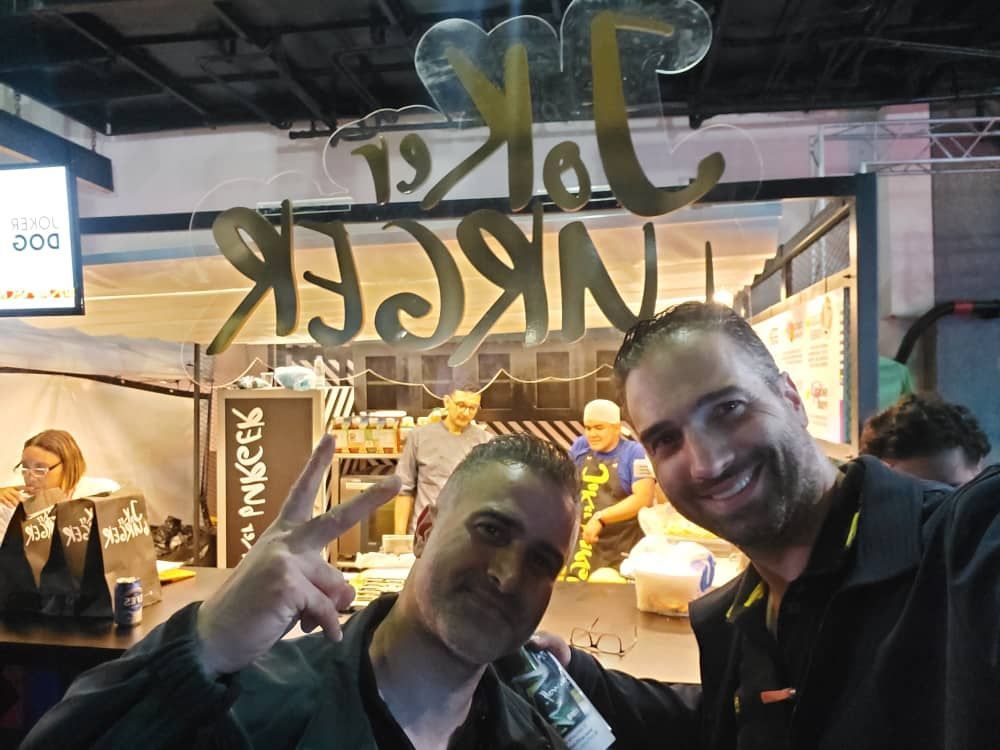 Two men smiling, one making a peace sign, in front of a mirrored food stall with the name "TAKE A NGER".