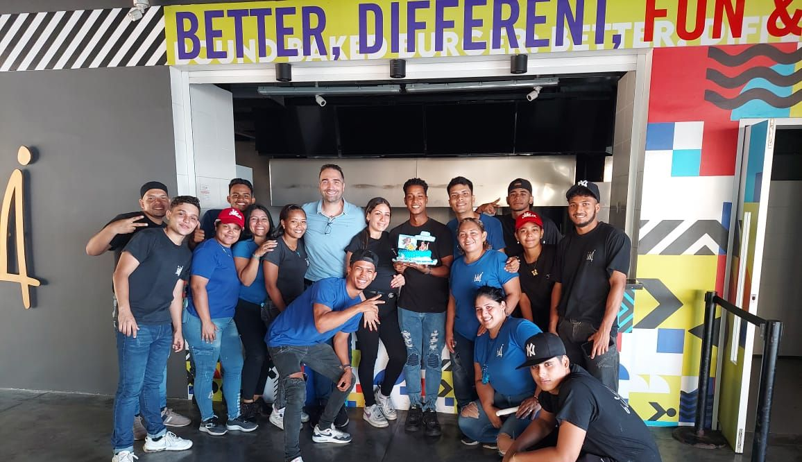 Group of people posing in front of a colorful wall; some wearing blue shirts and caps, holding a cake.
