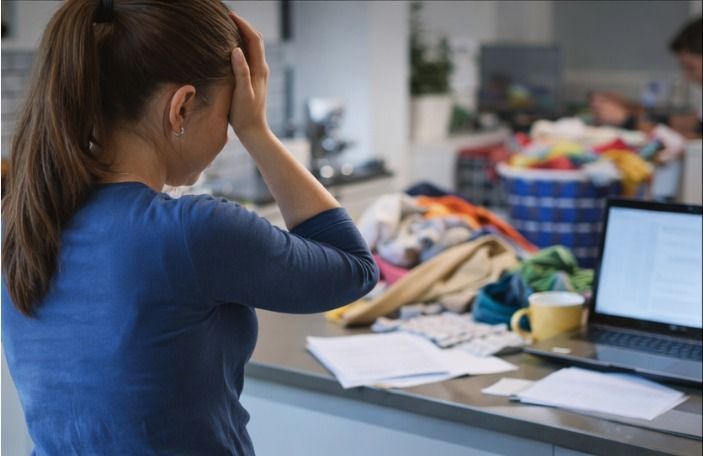 Woman with hands on head, overwhelmed by messy laundry, papers, and laptop on a kitchen counter.