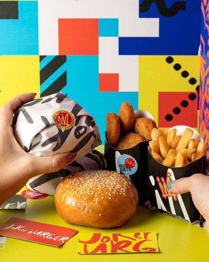 Hands holding a wrapped burger, fries, and nuggets with a sesame bun in front of a colorful backdrop.