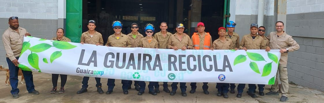 Group of people posing in front of a colorful wall; some wearing blue shirts and caps, holding a cake.