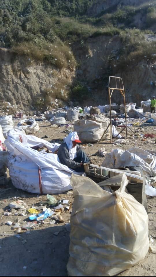 A person wearing a head covering sits among large bags and garbage at a waste site with a hillside backdrop.