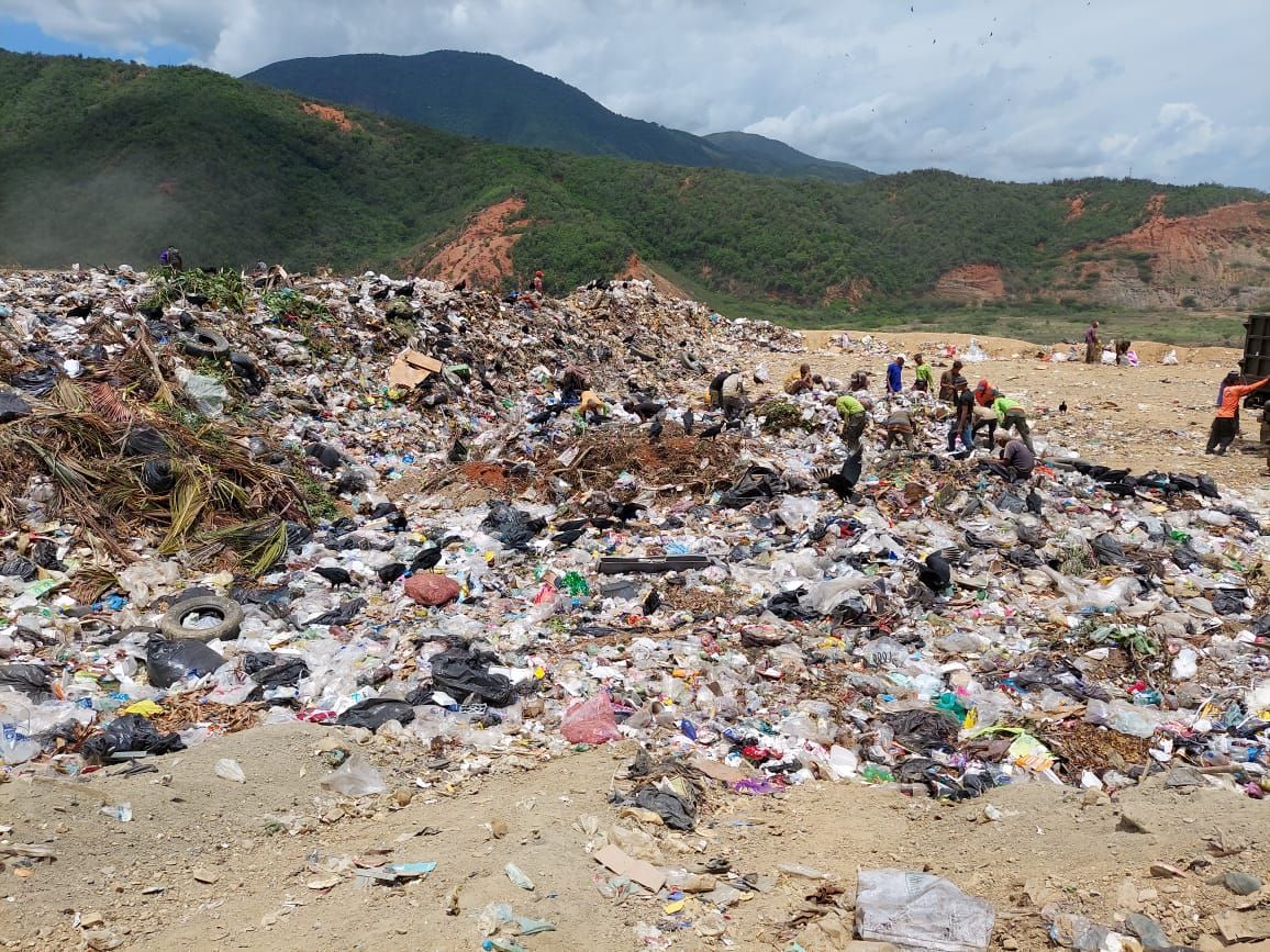 Group of people posing in front of a colorful wall; some wearing blue shirts and caps, holding a cake.
