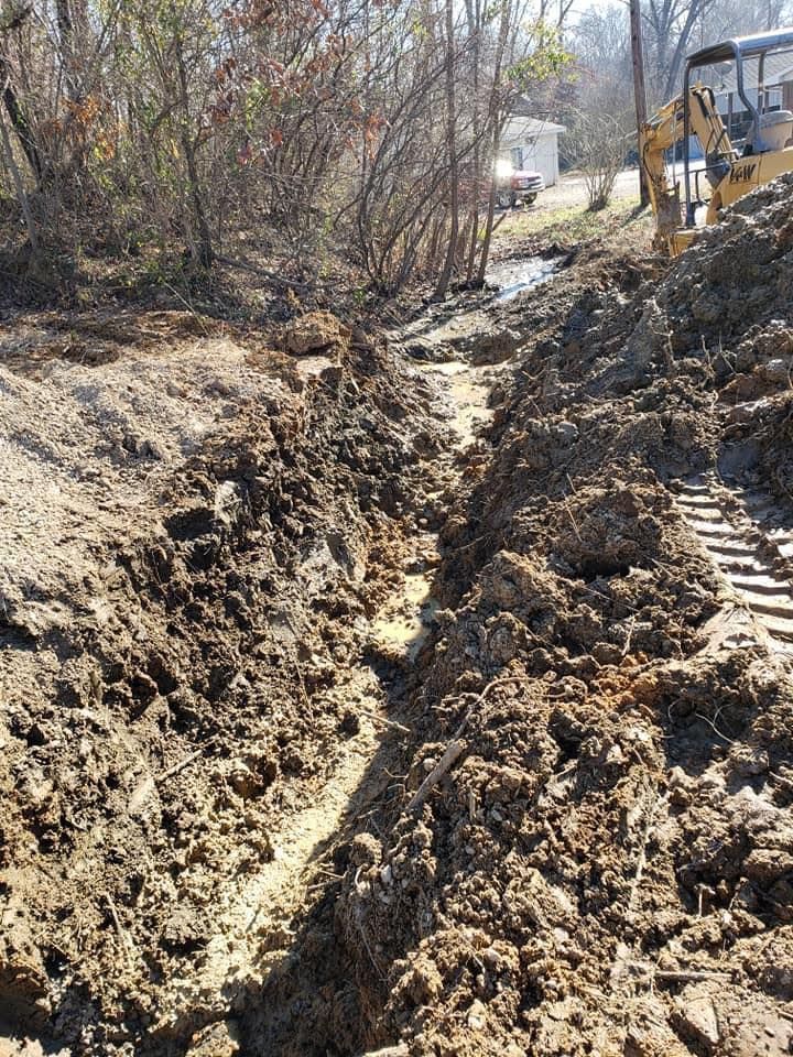Muddy trench dug through dirt and vegetation; excavator tracks visible.