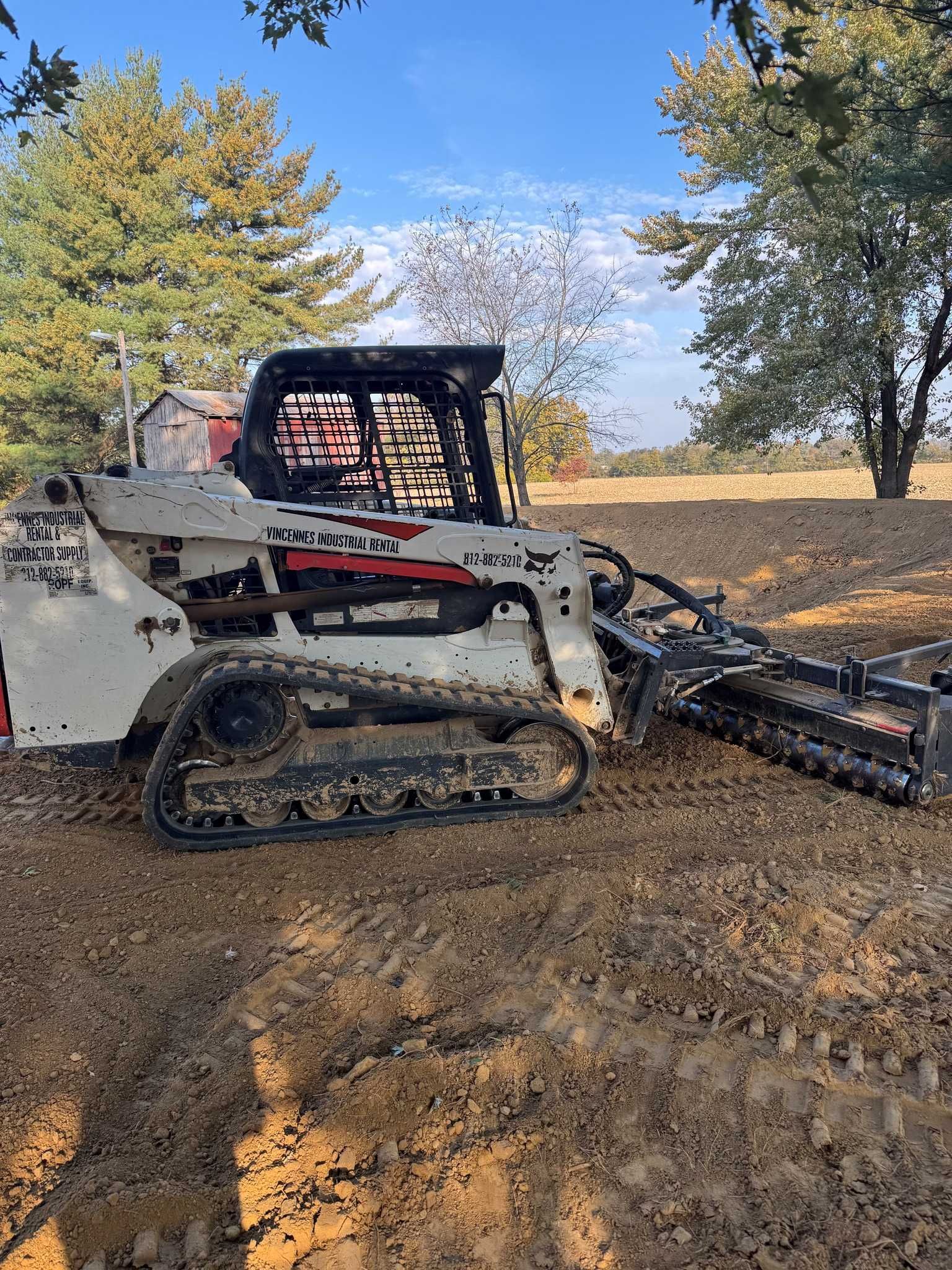 White Bobcat track loader with trencher attachment digging in a dirt field.