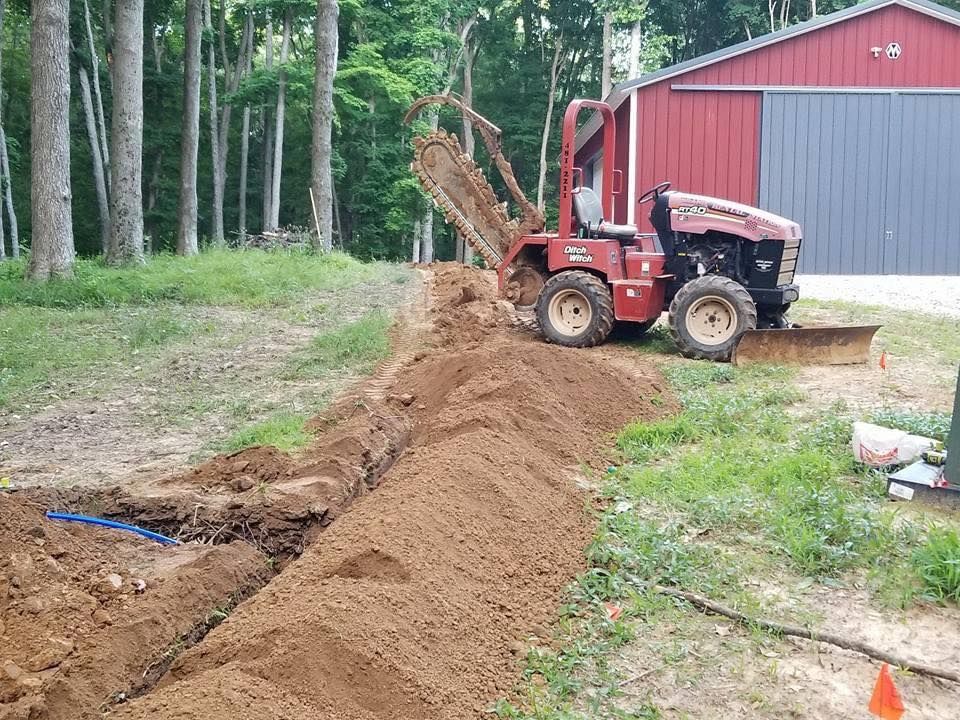A red trencher digs a trench in a grassy field. A barn is in the background.