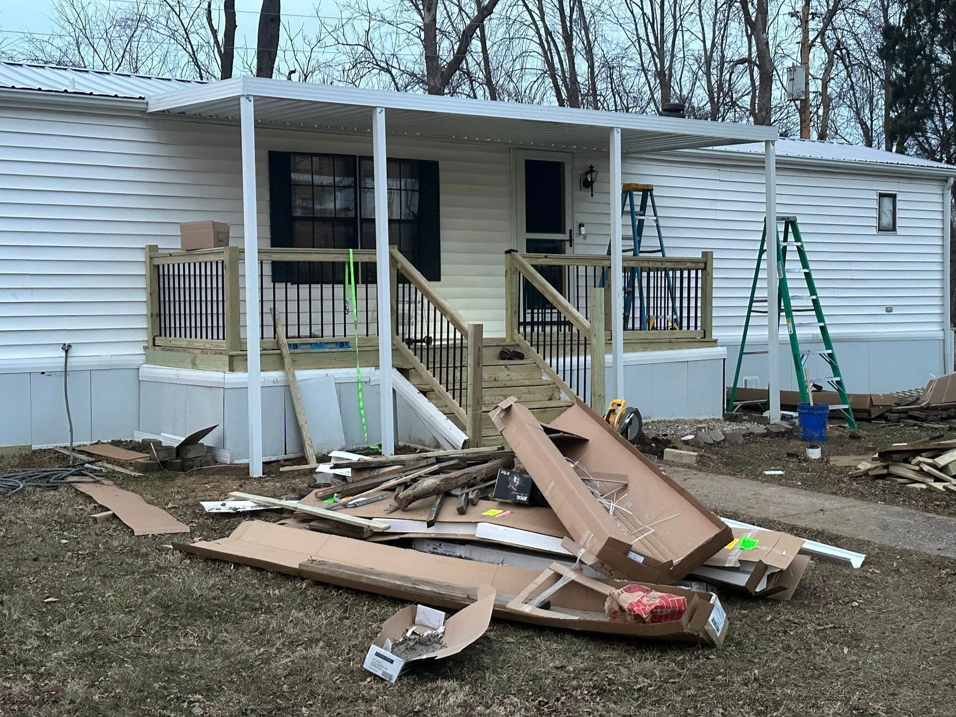 Mobile home with porch under construction, surrounded by building materials, and a ladder.