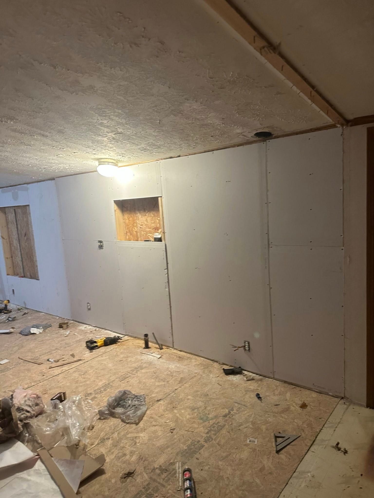 Interior wall under construction with drywall installed. Room has unfinished ceiling and floor, tools scattered.