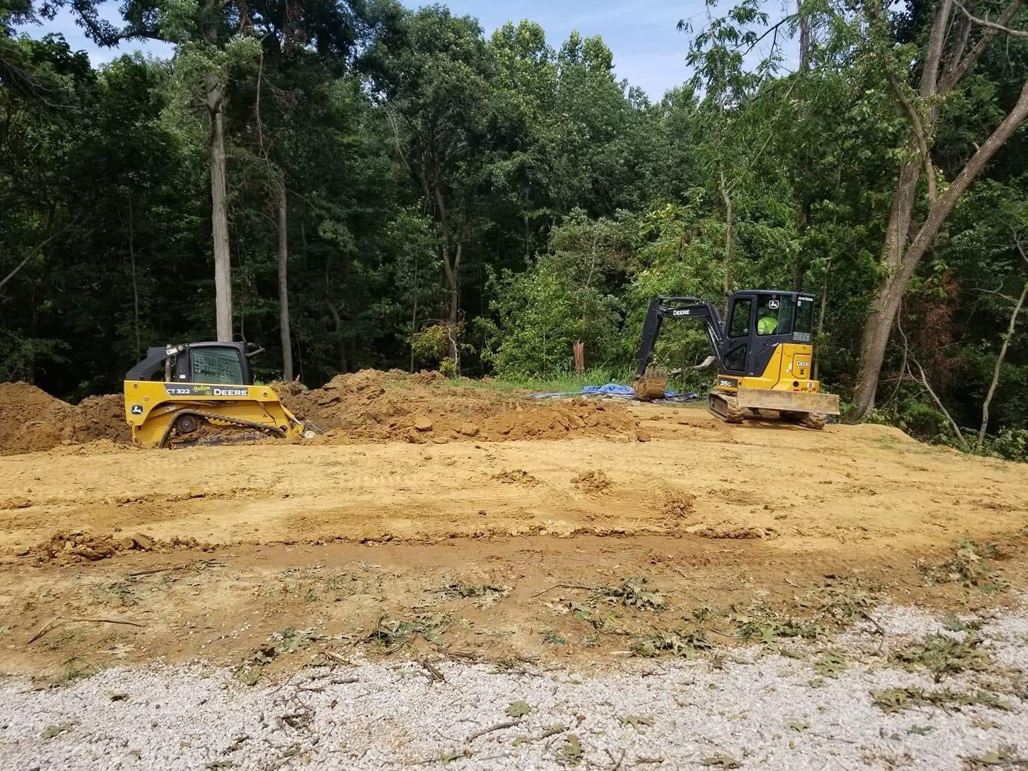 Two yellow construction vehicles on a dirt clearing in front of a treeline, under a bright sky.