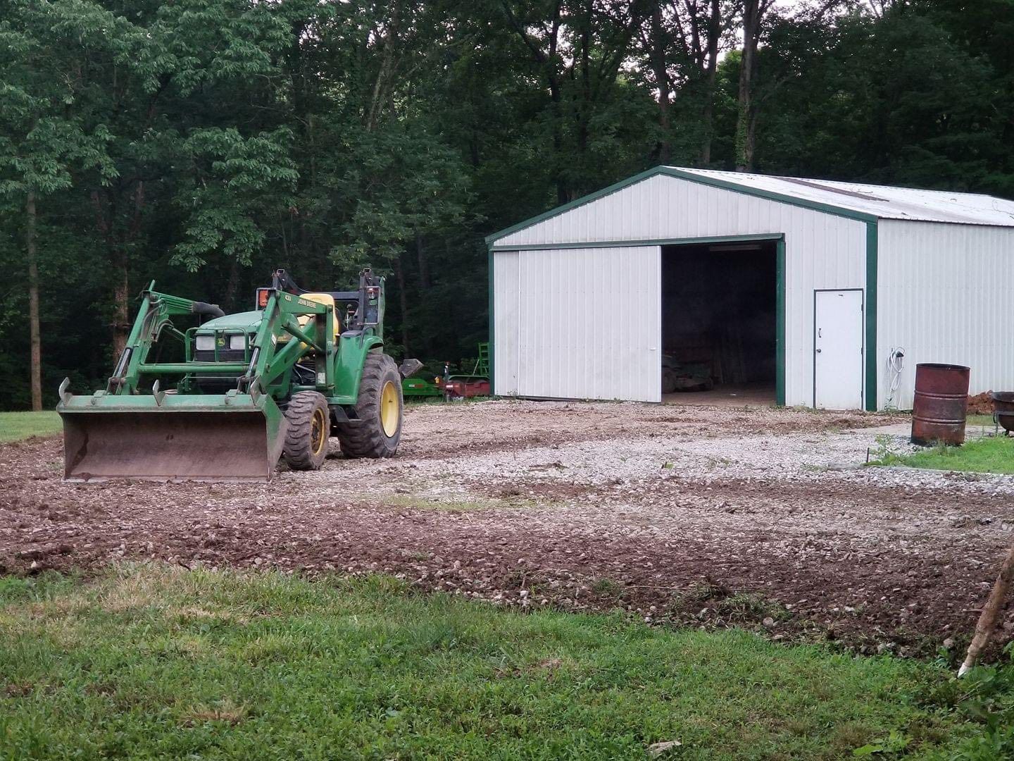 John Deere tractor grading gravel in front of a white metal shed with green trim.