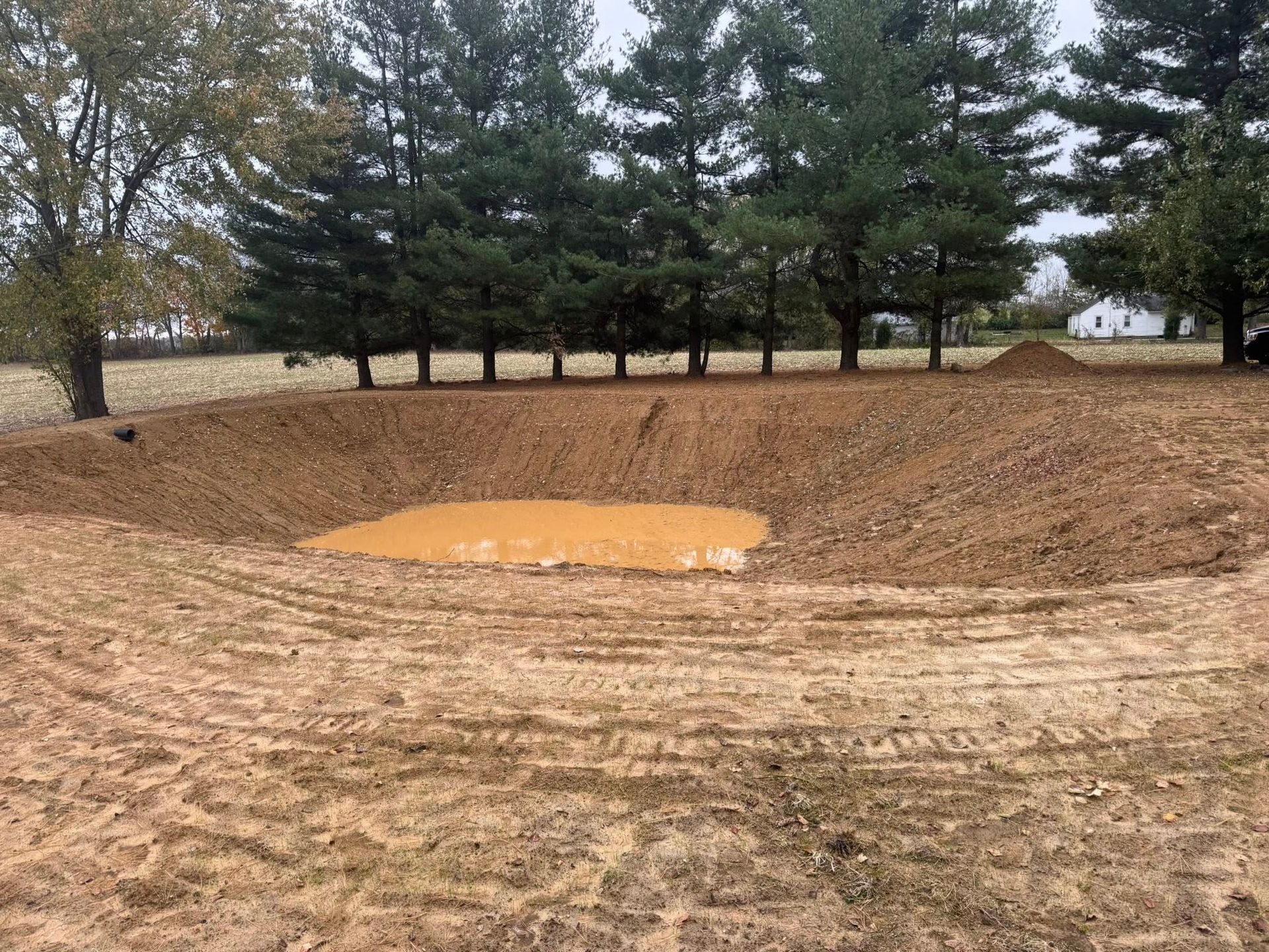 Newly dug pond with muddy water, surrounded by dirt and grass, trees in the background.