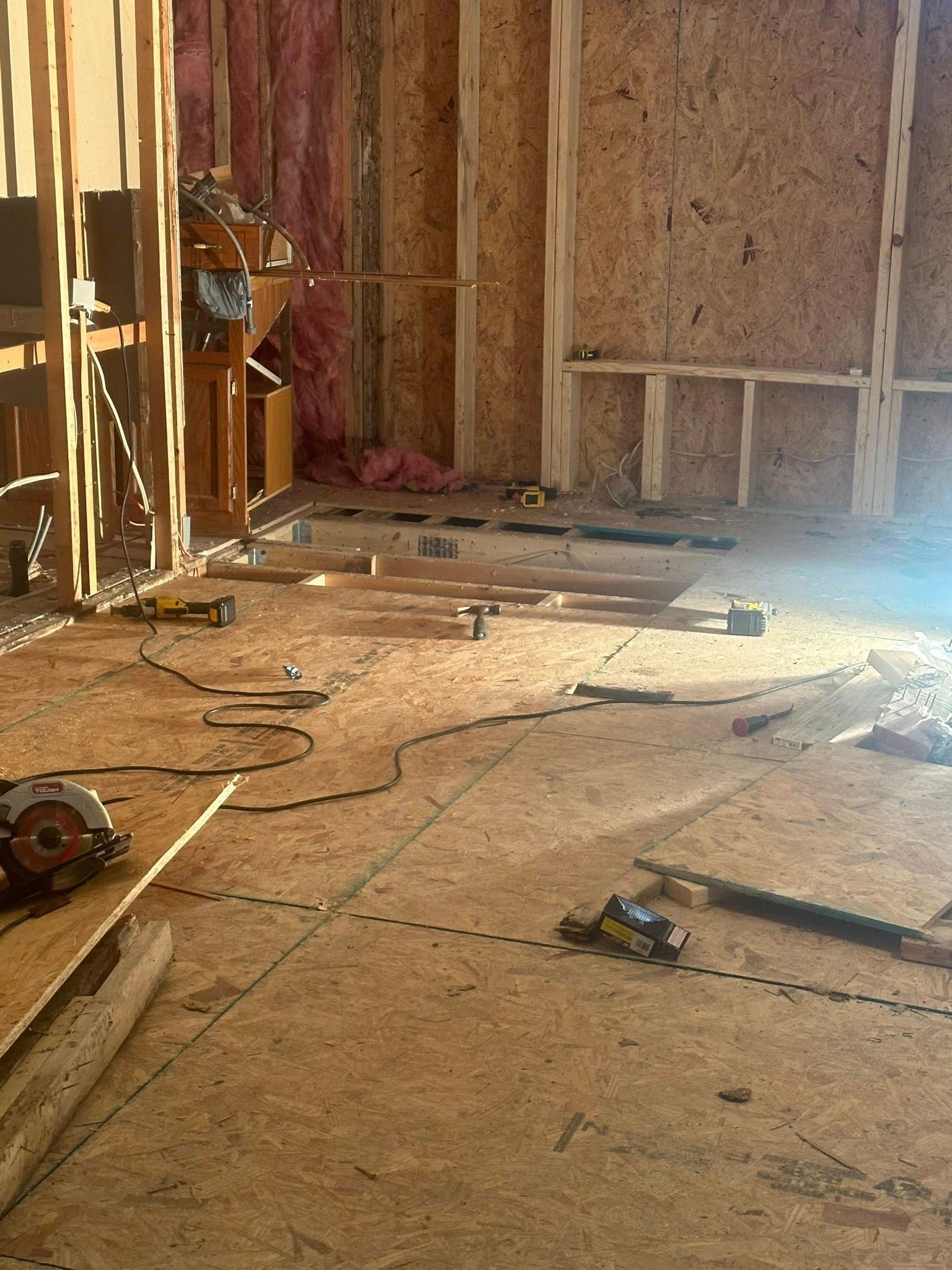 Construction site interior with plywood floor and wood framing. Tools and materials scattered around.