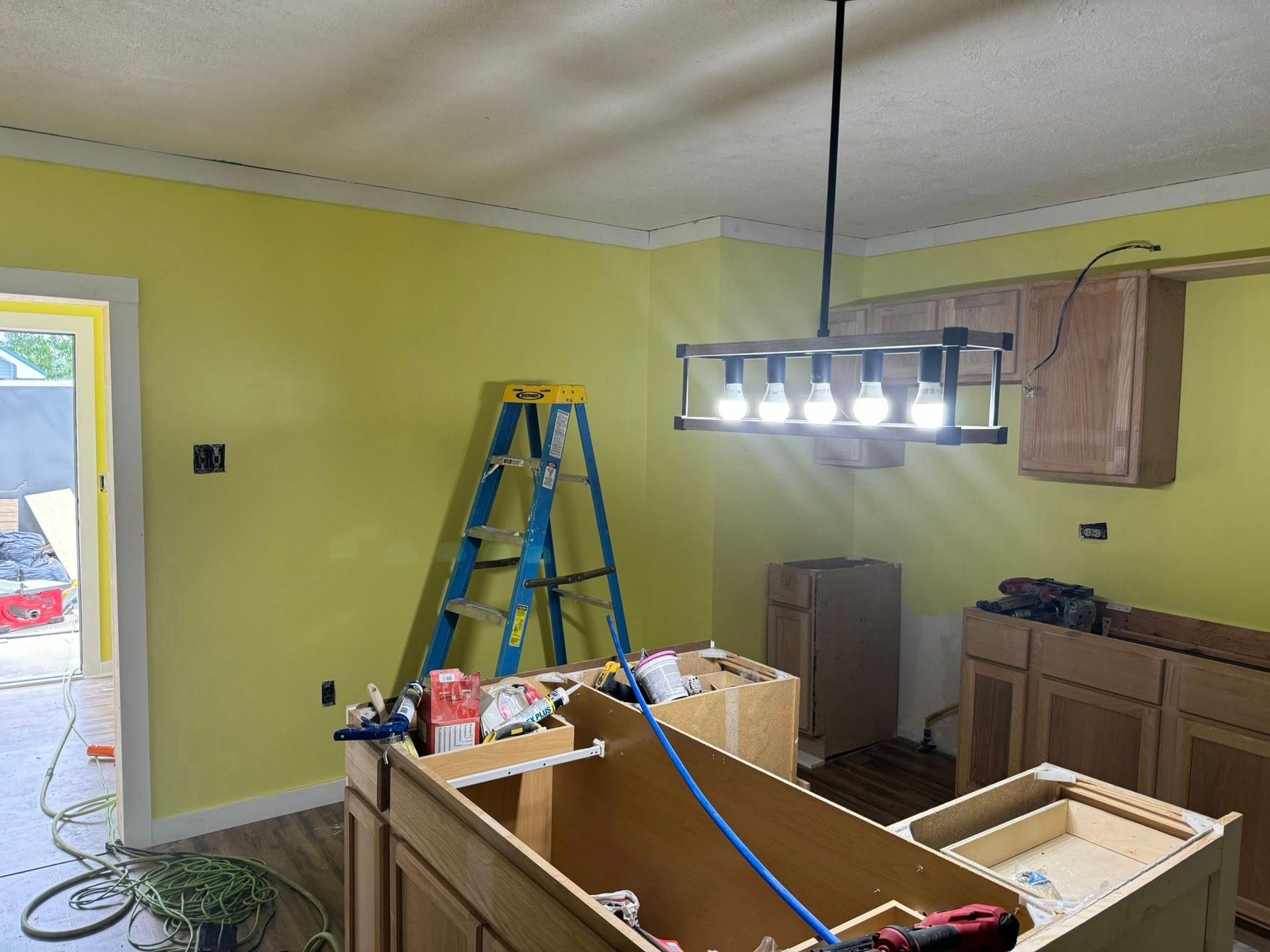 Kitchen undergoing renovation; yellow walls, hanging light fixture, cabinets, ladder.