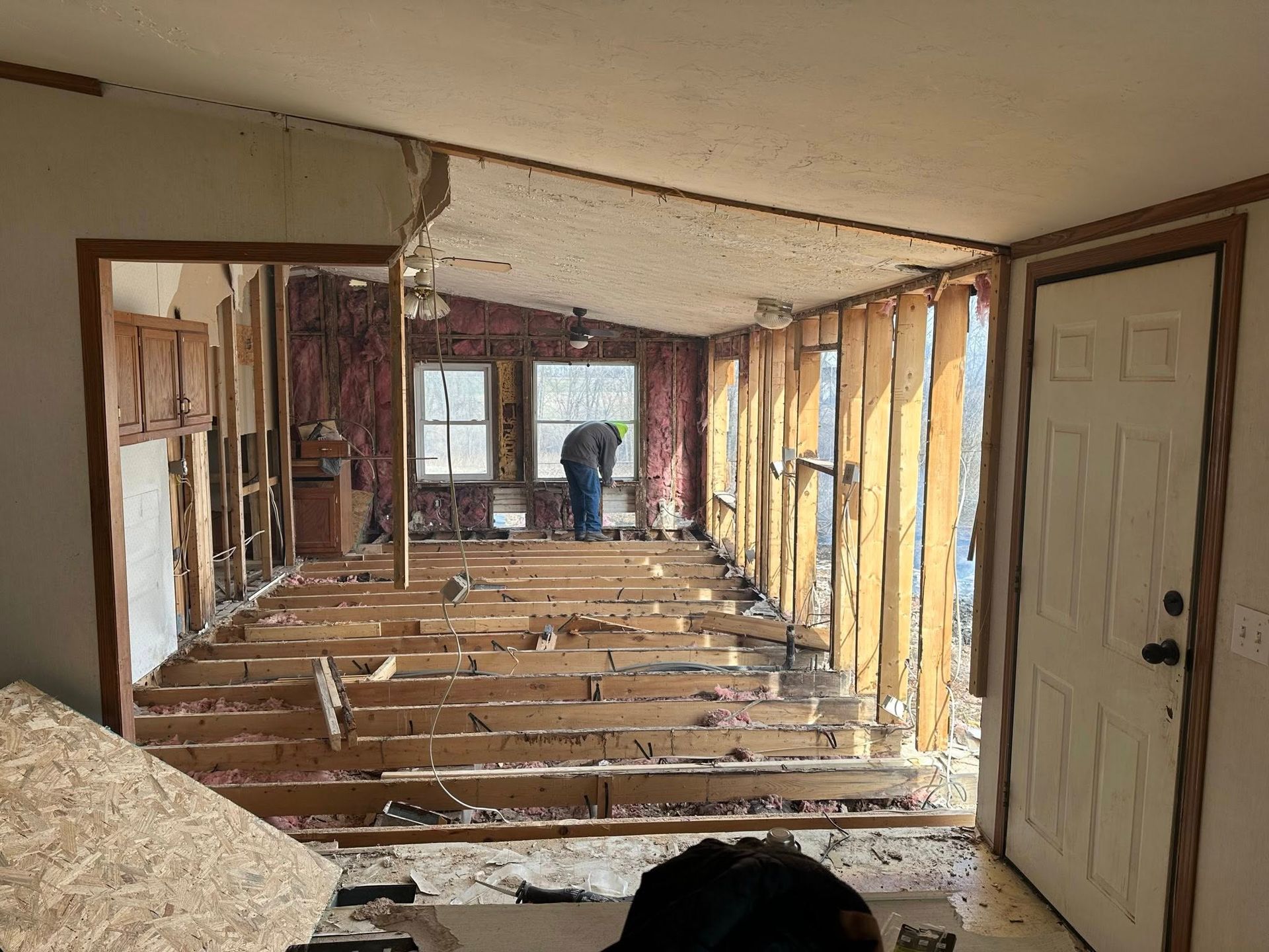 Interior home undergoing renovation: exposed floorboards, walls. Person works near windows. Doorway and door visible.