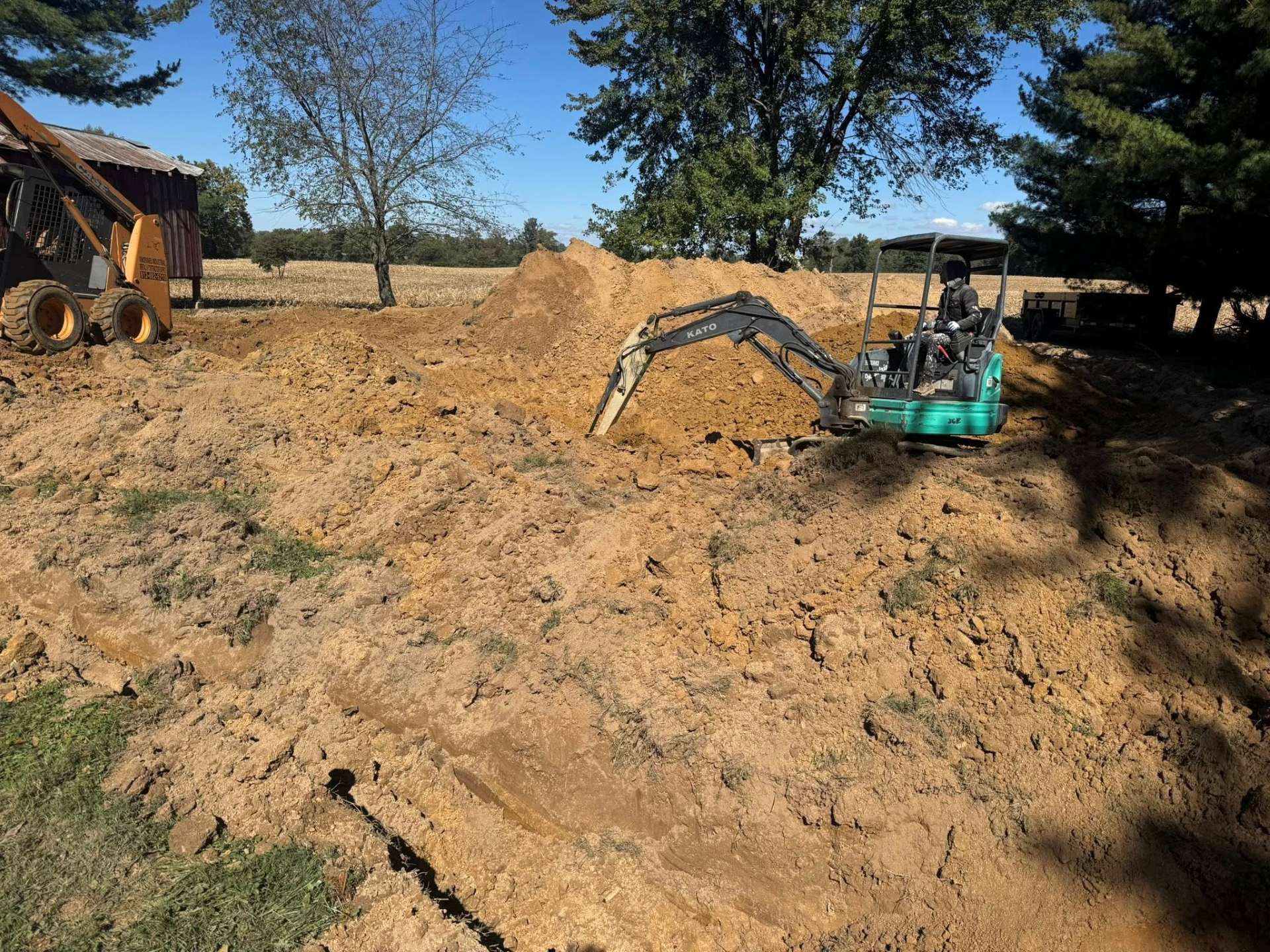 A small excavator digs into a pile of dirt in a field, with trees and a building visible in the background.