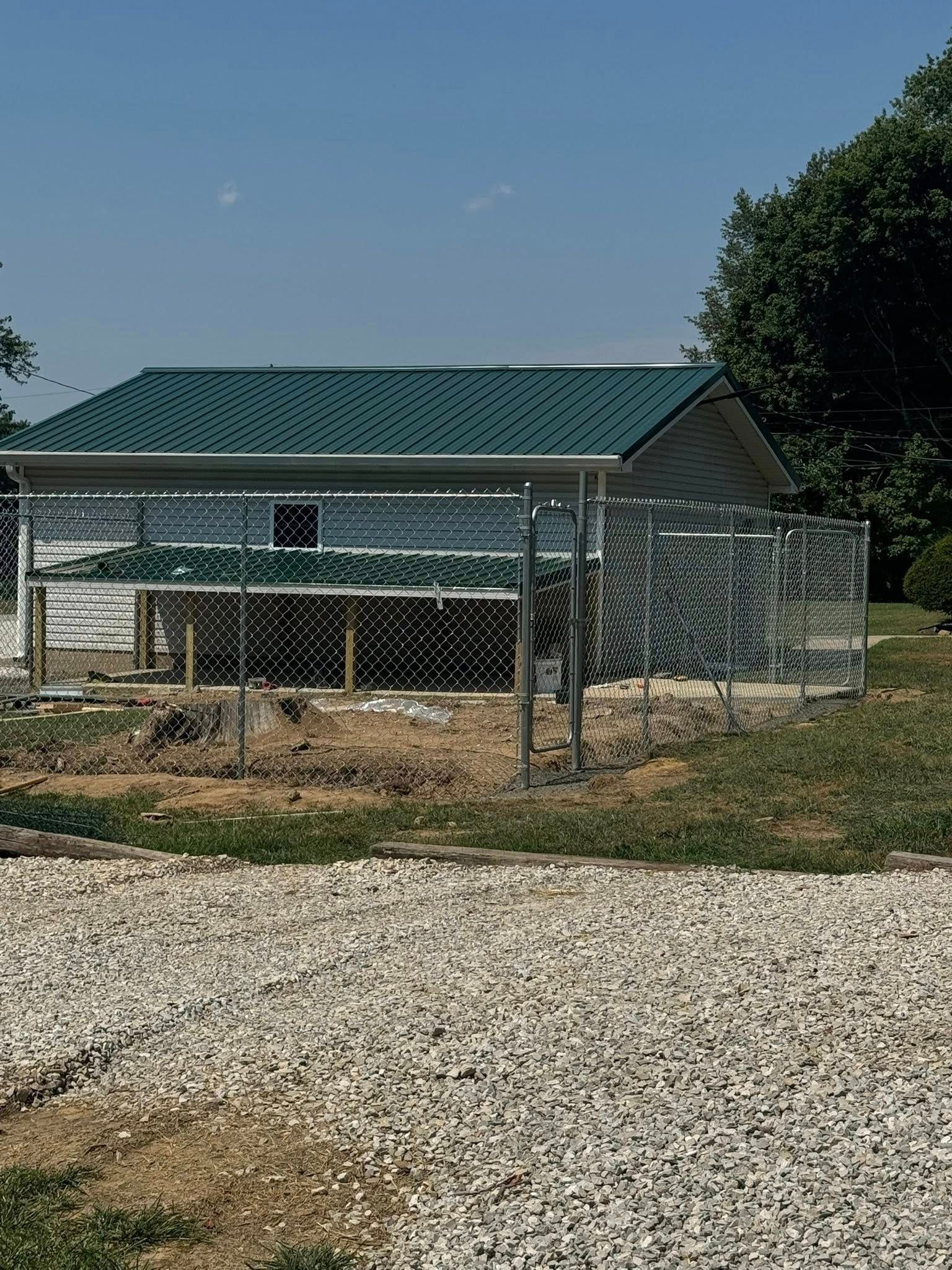 Chain-link fenced dog kennel with a green roof, set in a yard with gravel and grass.