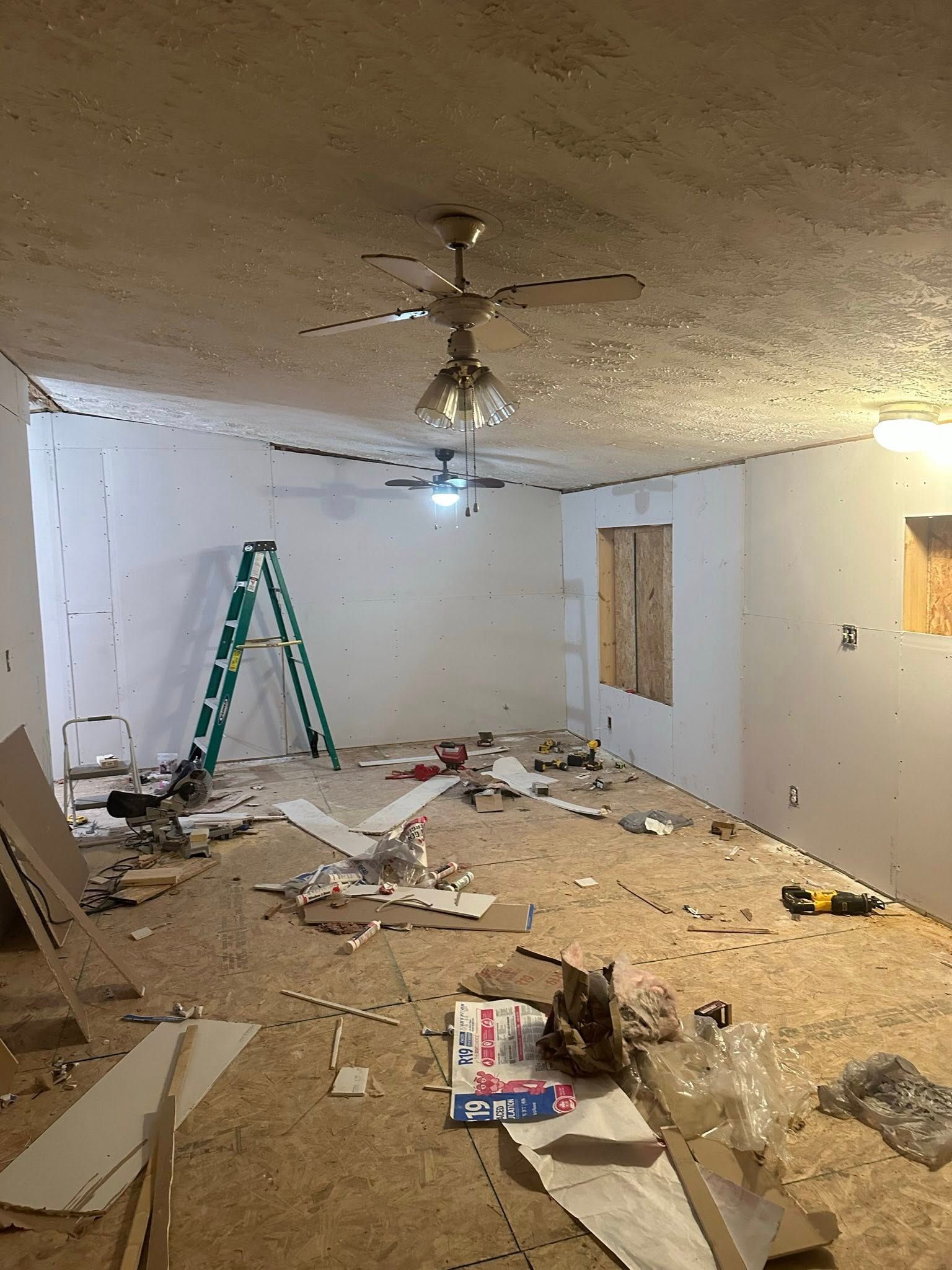Room under renovation with drywall, plywood floor, ladder, and ceiling fans.