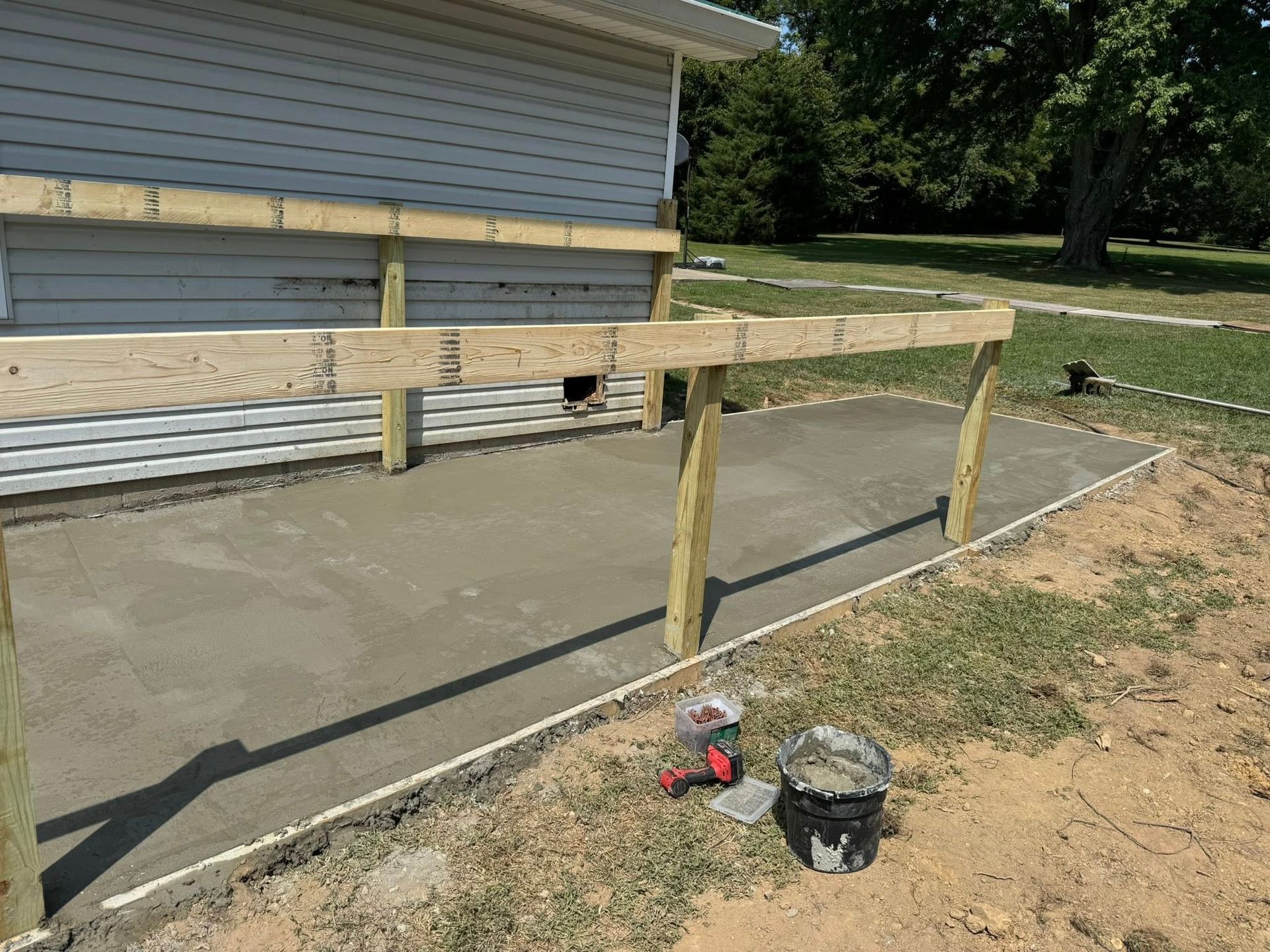 Concrete patio with wood supports and a building's siding in the background, set in a grassy yard.