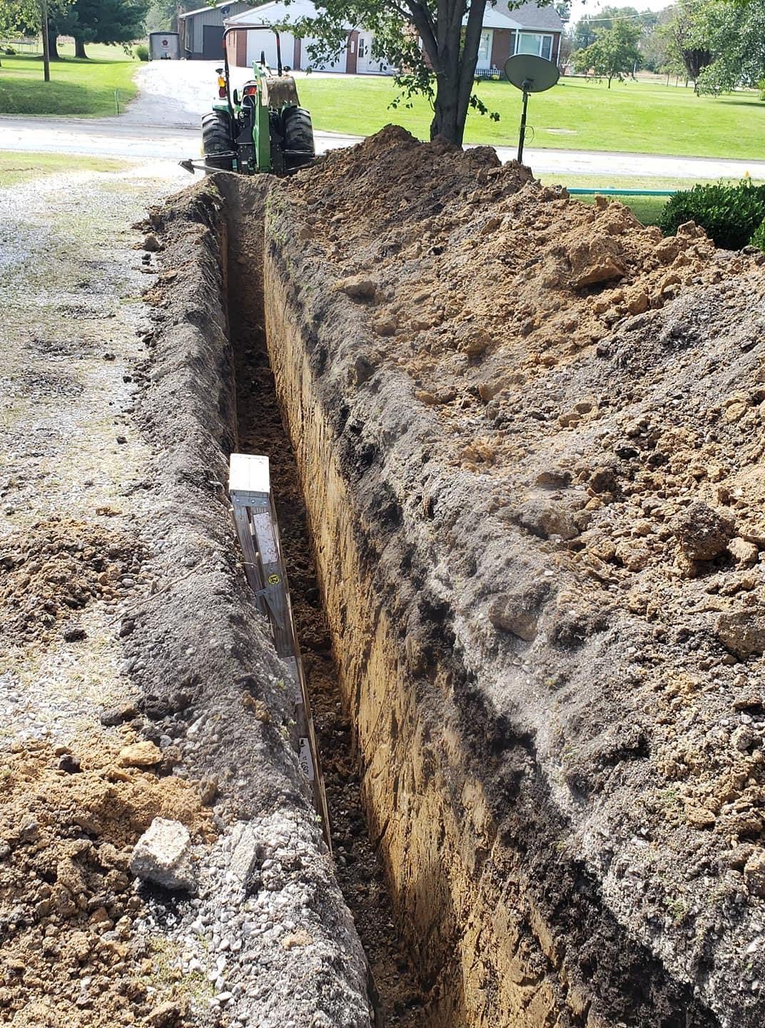 Long trench dug in the ground with a tractor in the background; dirt piles on the sides.