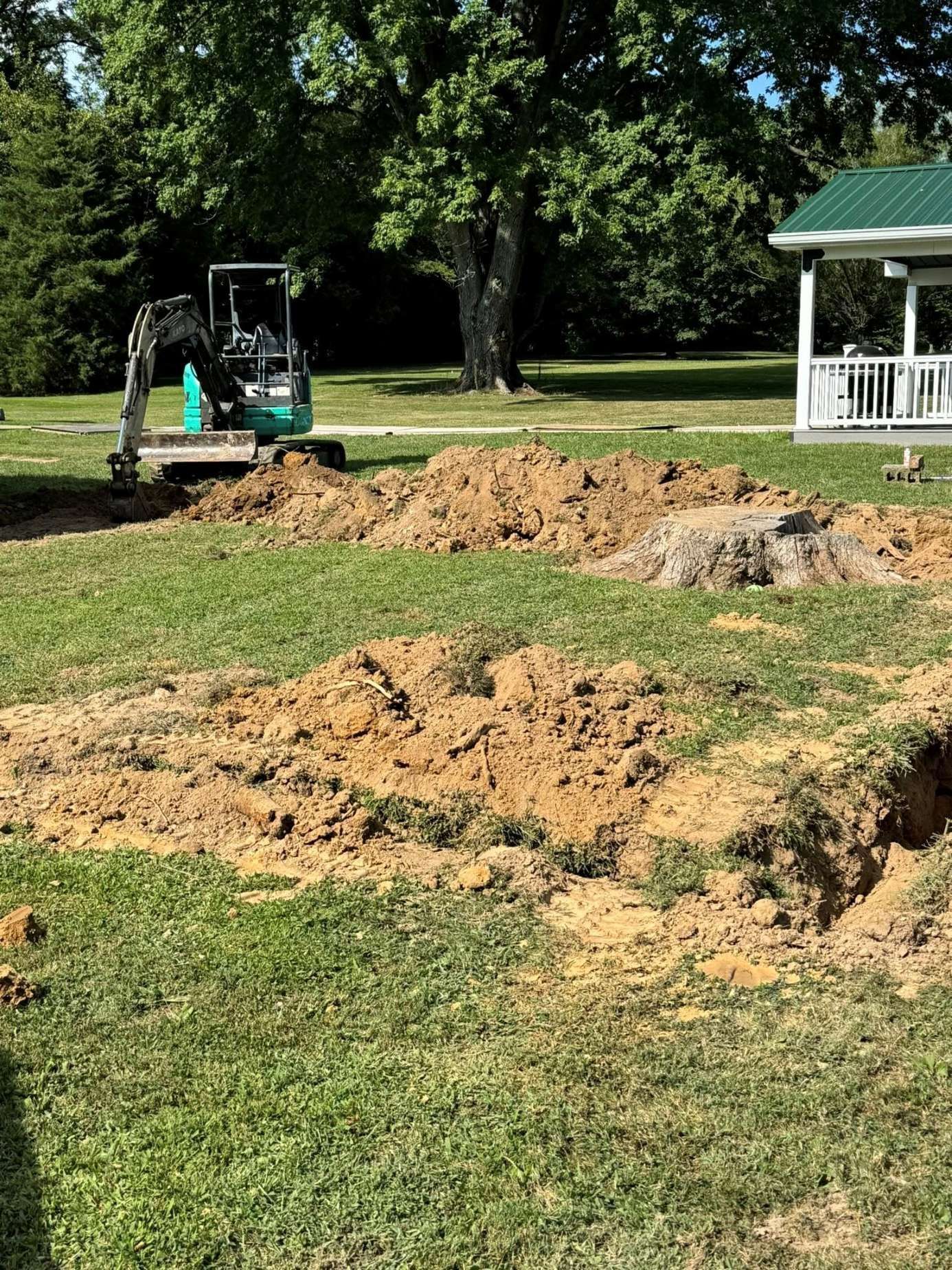 Mini-excavator digging in a grassy yard, piles of dirt nearby. A white gazebo and trees are in the background.