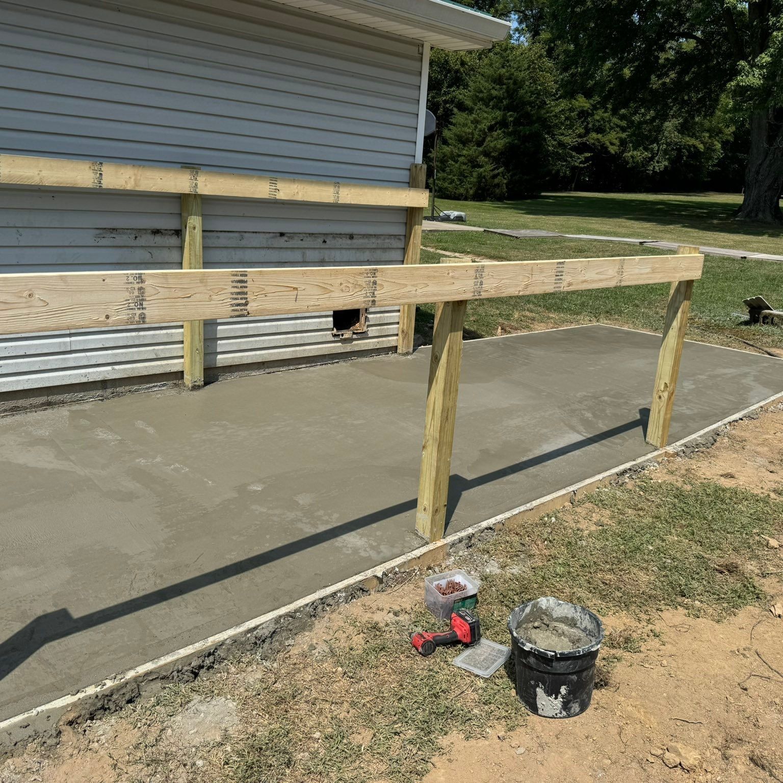 Concrete patio under construction with wooden frame, next to a building. Tools and materials are nearby.