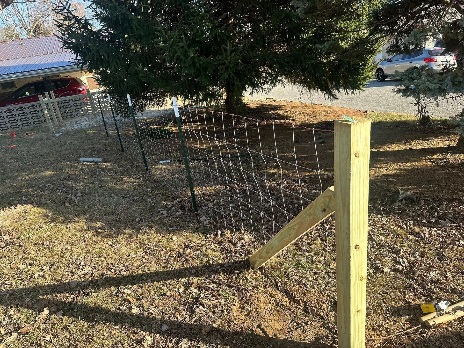 A partially built wooden fence with mesh, in a yard with a tree and a car visible.