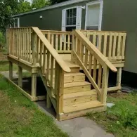 A new, light-colored wooden deck with stairs and railings attached to the side of a green mobile home.