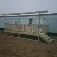 A new wooden deck with railings and a top support beam, built in front of a light-colored mobile home.