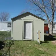A light green storage shed with a white door stands in a grassy yard under a clear blue sky, next to a mobile home.