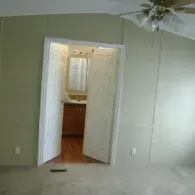 An open doorway reveals a bathroom with wooden cabinets, viewed from a room with light green paneled walls and carpet.