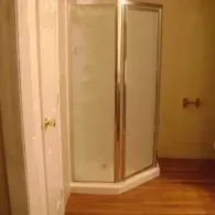 A corner shower stall with frosted glass panels and a silver frame sits on a wooden floor next to a white door.