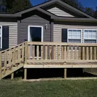 A new, light-colored wooden deck with stairs and a railing built in front of the entrance of a tan and grey house.