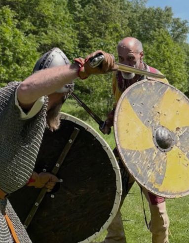 Two armored fighters with round shields and swords sparring outdoors in a grassy field.