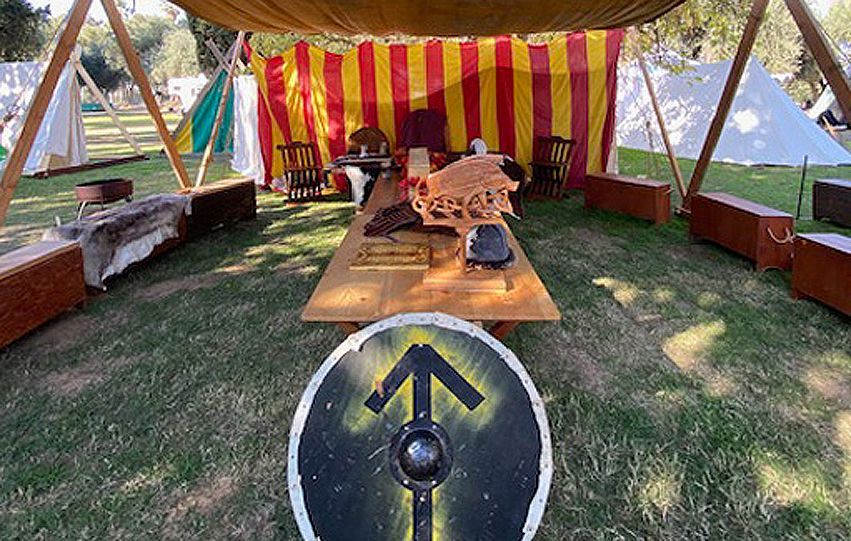 Striped tent campsite with wooden benches, fire pit, and a central table under shade trees