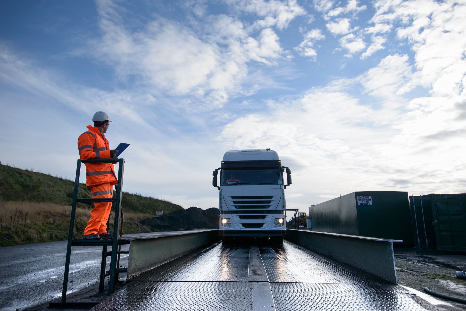 Worker checks truck on weighbridge under a partly cloudy sky.
