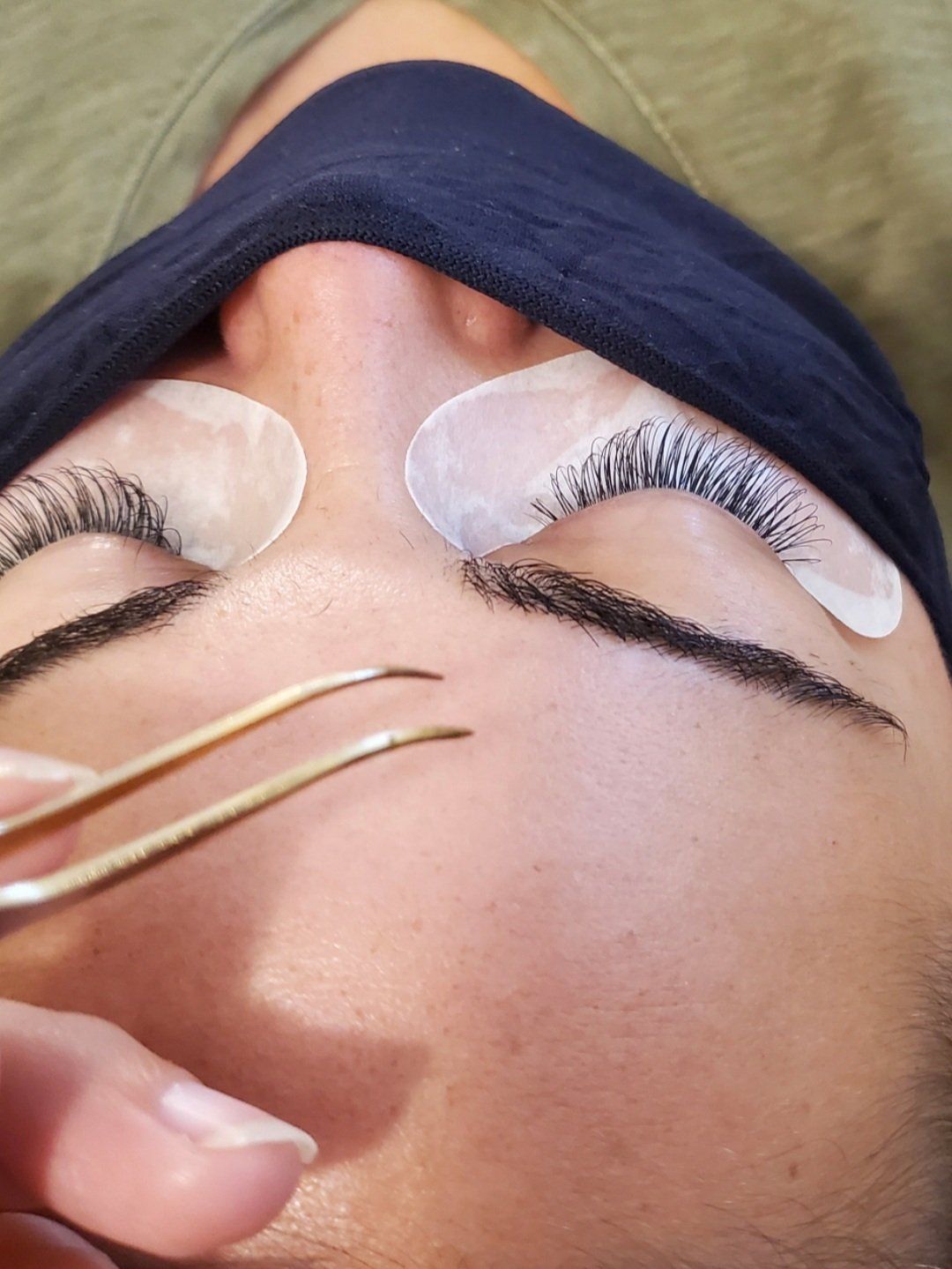 Person receiving eyelash extensions, eyes closed. Tweezers are held near the eyebrow.