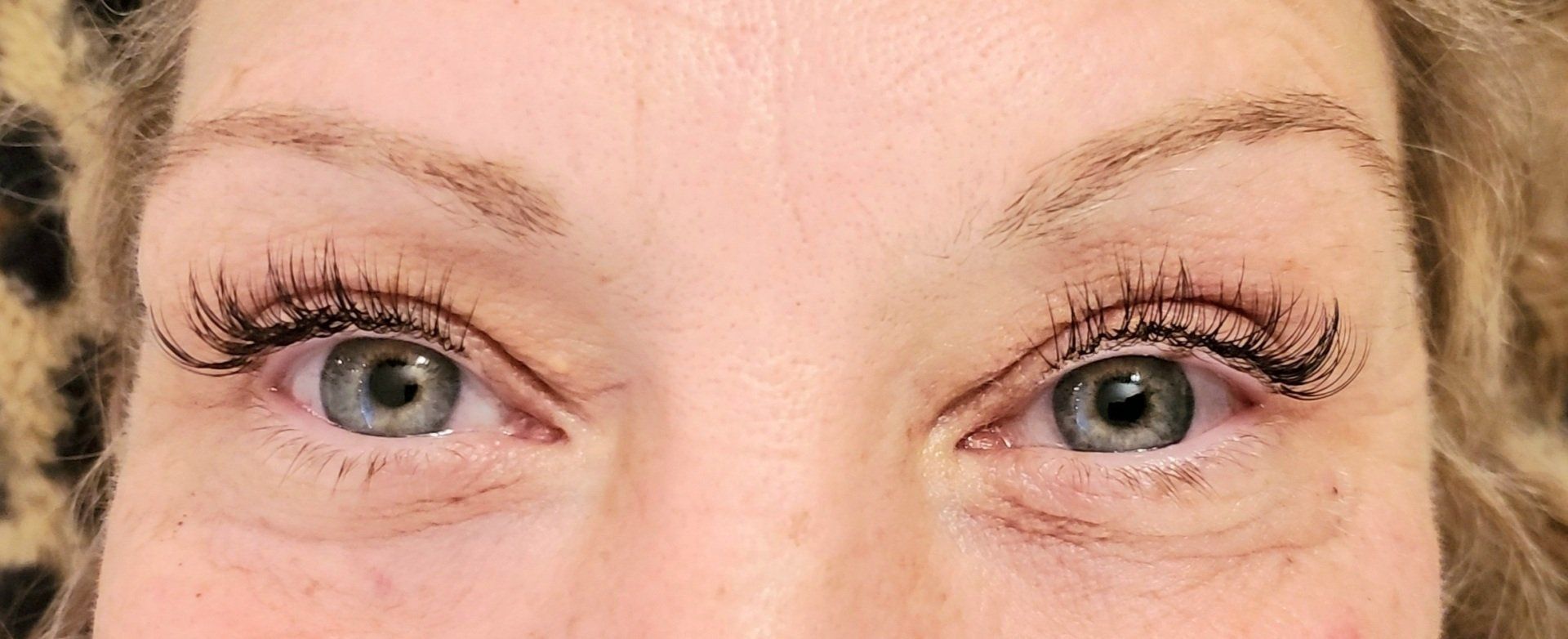 Close-up of a woman's eyes with long eyelashes, grey irises, and lightly filled-in eyebrows.