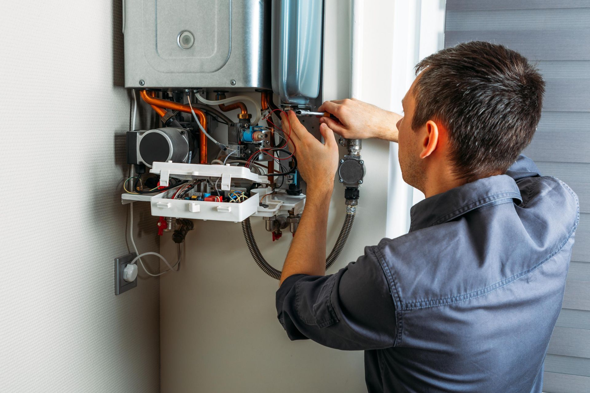 Person working on a water heater, indoors. Grey shirt, focused expression, using tools on the machine.
